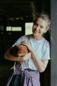Smiling young woman holding a chicken inside a rustic barn.