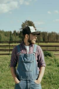 Bearded male farmer in denim overalls standing on a sunny day in a rural setting.